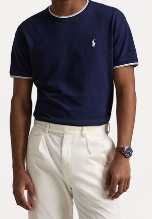 Marineblaues T-Shirt mit grauem Besatz, kurzen Ärmeln, mit kleinem weißem Logo. Kombiniert mit cremefarbenen Hosen und einer blauen Uhr.