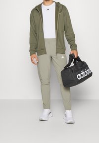 Sweat à capuche zippé vert olive avec bordure noire, t-shirt de sport blanc, pantalon gris clair fuselé, et sac de voyage noir avec un logo blanc. Baskets blanches.