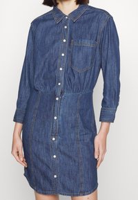 Džínové šaty tmavě modré barvy s dlouhými rukávy, límečkem, přední kapsou a knoflíkovým zapínáním uprostřed, s nařaseným pasem.