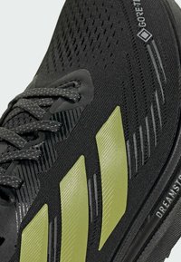 Black running shoe with olive green stripes, mesh upper, patterned laces, and "GORE-TEX" and "DREAMST" text on the side.