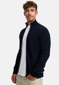 Chaqueta navy con cremallera, de patrón de punto texturizado, cuello alzado y cremallera contrastante. Lleva puesta sobre una camiseta blanca y pantalones de color claro.