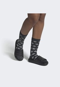 Chinelos pretos com três tiras texturizadas sobre meias cinzentas com padrões de folhas em preto e branco, usados em uma perna.