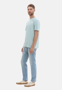 Helltürkises Baumwoll-T-Shirt mit kurzen Ärmeln und rundem Halsausschnitt, kombiniert mit hellen Denim-Jeans und cremefarbenen Sneakers mit braunen Sohlen.