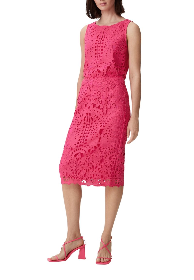 Robe en dentelle rose avec des découpes et motifs intriqués, présentant une silhouette ajustée et une jupe au genou, associée à des talons strappy assortis.