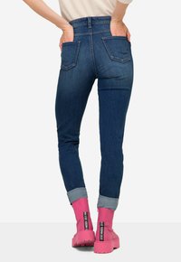 Tmavě modré slim džíny s vysokým pasem, pěti kapsami, mírně ohrnutými nohavicemi, v kombinaci s růžovými kotníkovými botami s výrazným brandingem.