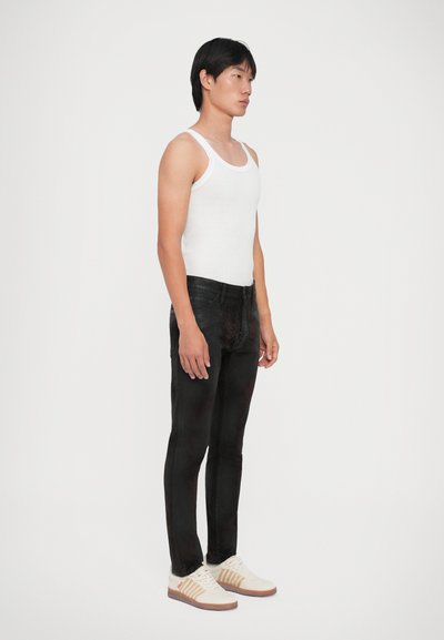 Canotta bianca a coste, jeans neri slim-fit e sneakers bianche con dettagli beige. Il modello è in piedi di lato contro uno sfondo neutro.