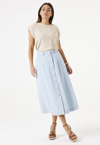 Lichtbeige top met korte mouwen, gecombineerd met een lichtblauwe midi-rok met knopen aan de voorkant in zachte denim. Model draagt bruine sandalen met bandjes.