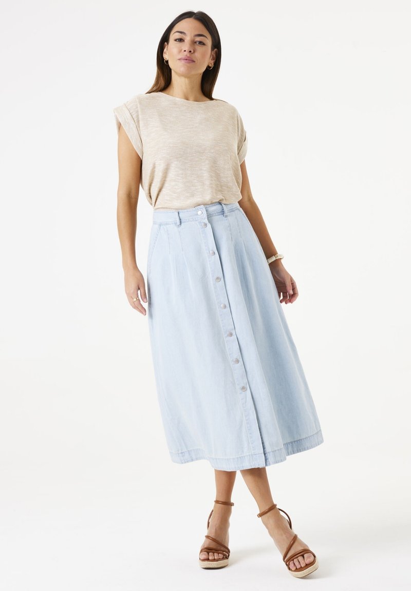 Lichtbeige top met korte mouwen, gecombineerd met een lichtblauwe midi-rok met knopen aan de voorkant in zachte denim. Model draagt bruine sandalen met bandjes.