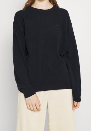 Kvinde iført sort langærmet trøje med diskret logo, kombineret med løstsiddende beige bukser, stående foran en ensfarvet baggrund.