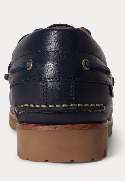 Granatowy skórzany mokasyn z brązową gumową podeszwą, przeszyciami w stylu mokasyna oraz ozdobnym sznurowaniem wokół pięty, widziany od tyłu.