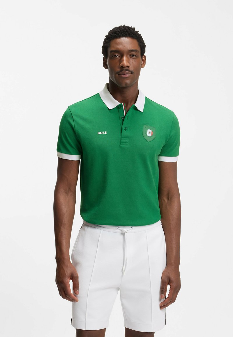 Homme portant un polo vert avec col et manches blanches, short blanc, debout devant un fond clair, faisant face à l'avant.