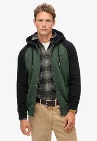 Sudadera con capucha verde y negra, con cierre frontal de cremallera, que presenta una textura suave y mangas en contraste, combinada con una camisa de franela a cuadros.