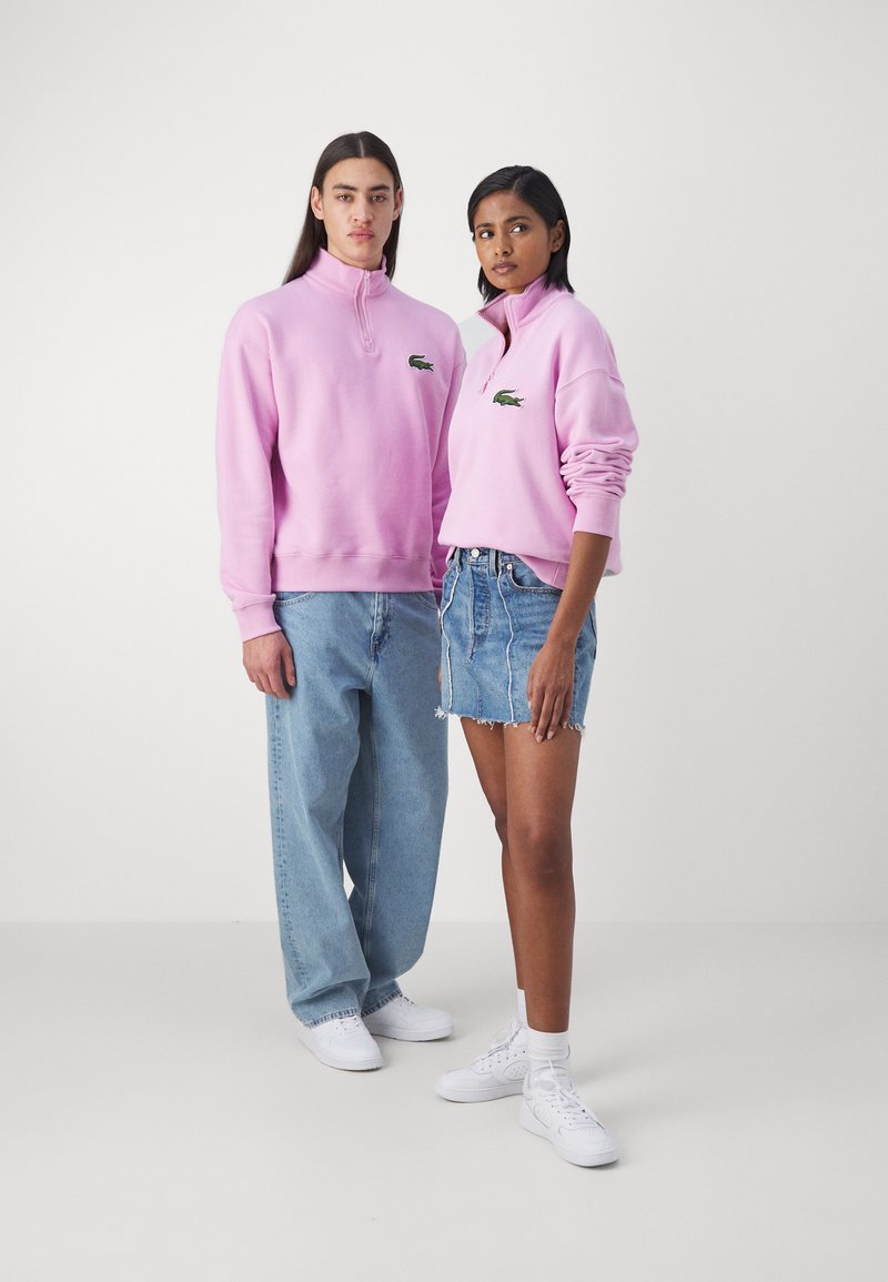 Due modelli indossano felpe leggere di colore rosa con colli a zip e loghi ricamati, abbinate a jeans azzurri e gonne di denim.