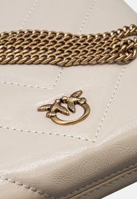 Borsa bianca in pelle trapuntata con catena dorata e chiusura decorativa con due uccelli. Texture liscia con dettagli di cucitura sottili.
