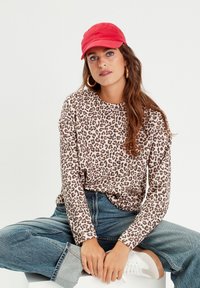 Camicia a maniche lunghe con stampa leopardata in beige e marrone, abbinata a jeans in denim a vita alta e un cappellino da baseball rosso. Sneakers bianche visibili.