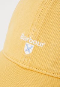 Sárga pamut sapka, texturált anyaggal, fehér hímzett márkanevével "Barbour" és egy kis pajzs logóval.