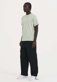 Camiseta de algodón verde claro con un pequeño logotipo blanco, combinada con pantalones cargo negros con bolsillos y bordados coloridos, usados con zapatillas blancas.