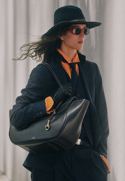 Mujer con sombrero negro de ala ancha, gafas de sol rojas, guantes negros, traje oscuro con camisa naranja, llevando un gran bolso negro al hombro.