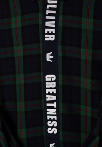 Koszula w kratę z ciemnoniebieskim, zielonym i czerwonym kolorem. Zawiera odważną białą wstążkę z napisem „WIELKOŚĆ” i grafiką korony.