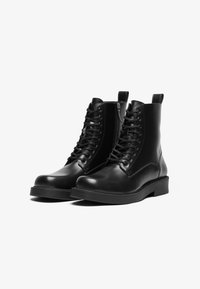 Botine din piele neagră până la gleznă cu sistem de șireturi în față, vârf rotund și talpă plată din cauciuc. Include un tab pentru a fi ușor de îmbrăcat.