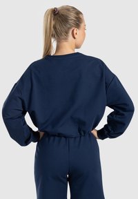 Felpa blu navy con vestibilità rilassata, spalle scese ed bordo elastico. Tessuto morbido con polsini a coste. La vista posteriore mostra un design minimal.