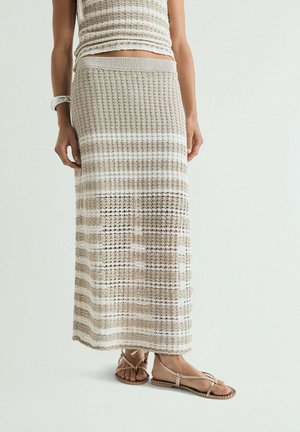 Mujer con falda maxi de punto a rayas beige y blanca, top sin mangas de punto a juego, pulsera metálica y sandalias planas beige con tiras.