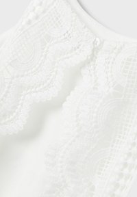 Weißer Stoff mit filigranen weißen Spitzenverzierungen am Ausschnitt, die kreisförmige Muster und Pompon-Akzente entlang der Kanten aufweisen.