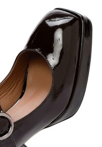 Zwarte gepatineerde leren plateau schoen met een ronde neuzen, open zijkant en cirkelvormige zilveren hardware detail. Een gladde textuur en contrasterend tan binnenzool.