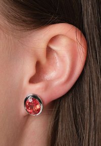 Rote Edelstein-Studiobohrringe mit einer silbernen Fassung, ausgestattet mit einem rund geschliffenen Stein und einer polierten Metalloberfläche, getragen vor einem hellen Haarhintergrund.