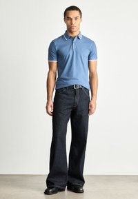Polo shirt bleu clair avec des détails blancs, associé à un jean évasé bleu foncé et des chaussures noires, posé devant un fond neutre.