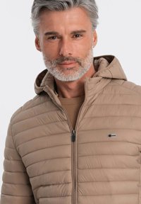 Beige pufferjakke med vandret syninger, hætte og lynlås. Har et lille mærke logo på venstre bryst. Glat, letvægtsmateriale.