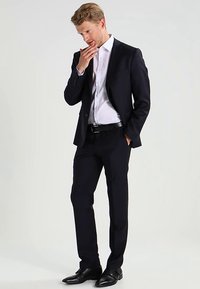 Abito blu navy dal design strutturato, con rever a lancia, camicia bianca elegante e scarpe di pelle nere; tessuto liscio con vestibilità su misura.