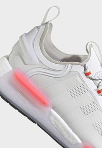 Vit träningsskor med ovandel i mesh, tre horisontella ränder och en genomskinlig sula. Anmärkningsvärda detaljer inkluderar röda accenter och en strukturerad häl.