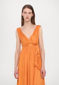 Orange omslagsklänning med djup V-ringning, texturerad tyg och knytning i midjan. Har mjuka plisseringar och är ärmlös.