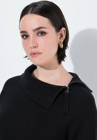 Zwarte sweater met een brede kraag, zijritsdetail en textuurstof. Model draagt gouden hoepel oorbellen en heeft halflang haar.