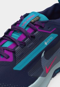 Κοντινό πλάνο μπλε και μοβ παπουτσιού τρεξίματος Nike Pegasus Trail με ροζ λεπτομέρειες, ετικέτα Gore-Tex, κορδόνια με σχέδιο και ορατή υφή στη σόλα.