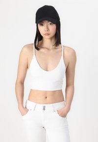 White cropped tank top with thin straps, paired with high-waisted white jeans featuring a button closure. A black baseball cap completes the look.