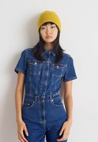Denim jumpsuit in medium blue met korte mouwen, twee voorzakken en een getailleerde taille. Draagt een geribbelde mosterdgele beanie.