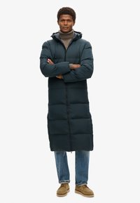 Lang pufferjack in donkergroen, met een capuchon, volledige rits en gewatteerd ontwerp. Gecombineerd met lichtblauwe jeans en beige schoenen.