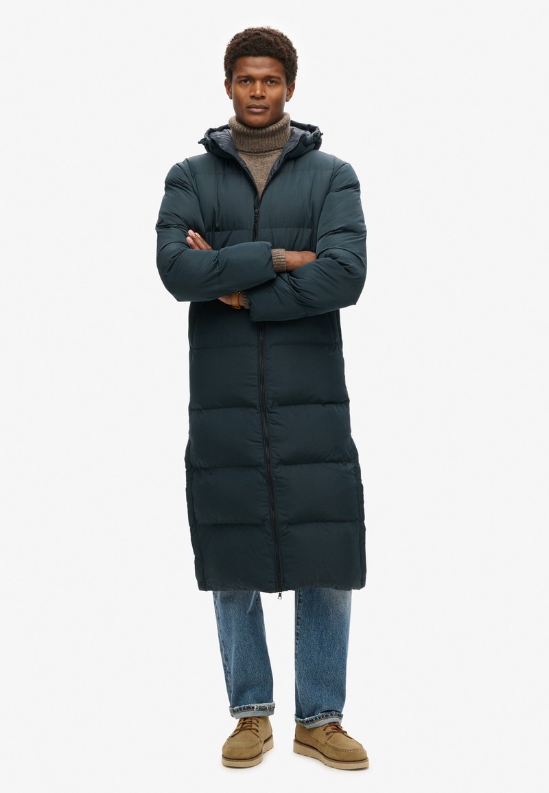 Lang pufferjack in donkergroen, met een capuchon, volledige rits en gewatteerd ontwerp. Gecombineerd met lichtblauwe jeans en beige schoenen.