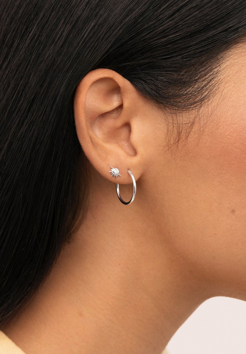 Orecchino a cerchio in argento con un accento a raggi di sole che presenta una pietra trasparente. Il design combina forme lisce e rotonde con una finitura lucida.