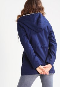 Giacca a vento blu navy con cappuccio e cordoncino, dotata di un rivestimento in pelliccia sintetica, tasche a patta e una vestibilità sartoriale. Tessuto morbido e testurizzato.