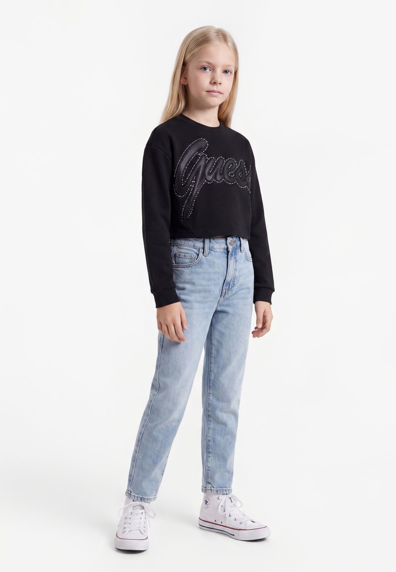 Jeune fille debout avec de longs cheveux blonds, portant un sweat Guess noir, un jean bleu clair et des baskets montantes blanches.