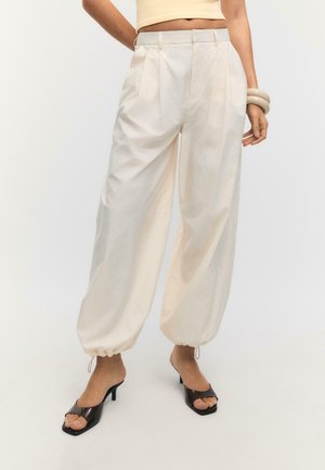 Žena oblečená v krémových voľných nohaviciach s vysokým pásom a stiahnutými lemami na šnúrky, čiernych sandáloch s otvorenou špičkou a bezrukávovom béžovom krátkom topu.