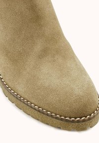 Bottine en daim beige clair avec un bout arrondi, une surface texturée et une semelle en caoutchouc ornée de surpiqûres visibles.