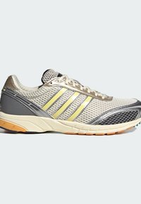 Αθλητικό παπούτσι σε ανοιχτό γκρι με επάνω μέρος από δίχτυ, τρεις κίτρινες ρίγες, αντανακλαστικές ασημένιες λεπτομέρειες, επένδυση στο κολάρο και υφή στην σόλα με πορτοκαλί καουτσούκ.