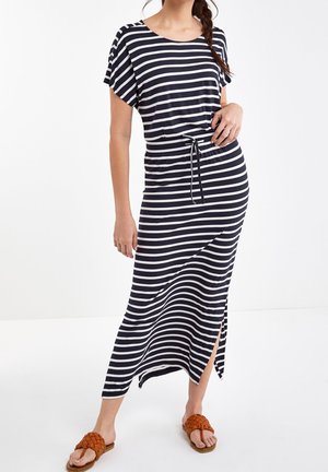 Femme portant une robe rayée noire et blanche à rayures horizontales avec une ceinture à la taille, une fente latérale, et des sandales plates tressées marron.