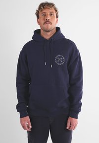 Hoodie bleu marine en tissu doux avec une poche avant, une capuche à cordon, et un logo circulaire sur la partie gauche de la poitrine.