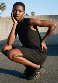 Fiatal nő fekete ujjatlan ruhában és tornacipőben guggol a járdán, egyik kezét a falnak támasztva, mögötte pálmafák.