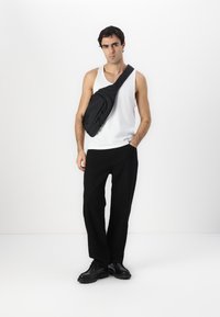 Sac bandoulière noir en tissu durable, avec un compartiment zippé et une bandoulière ajustable. Le modèle porte un débardeur blanc et un pantalon noir.